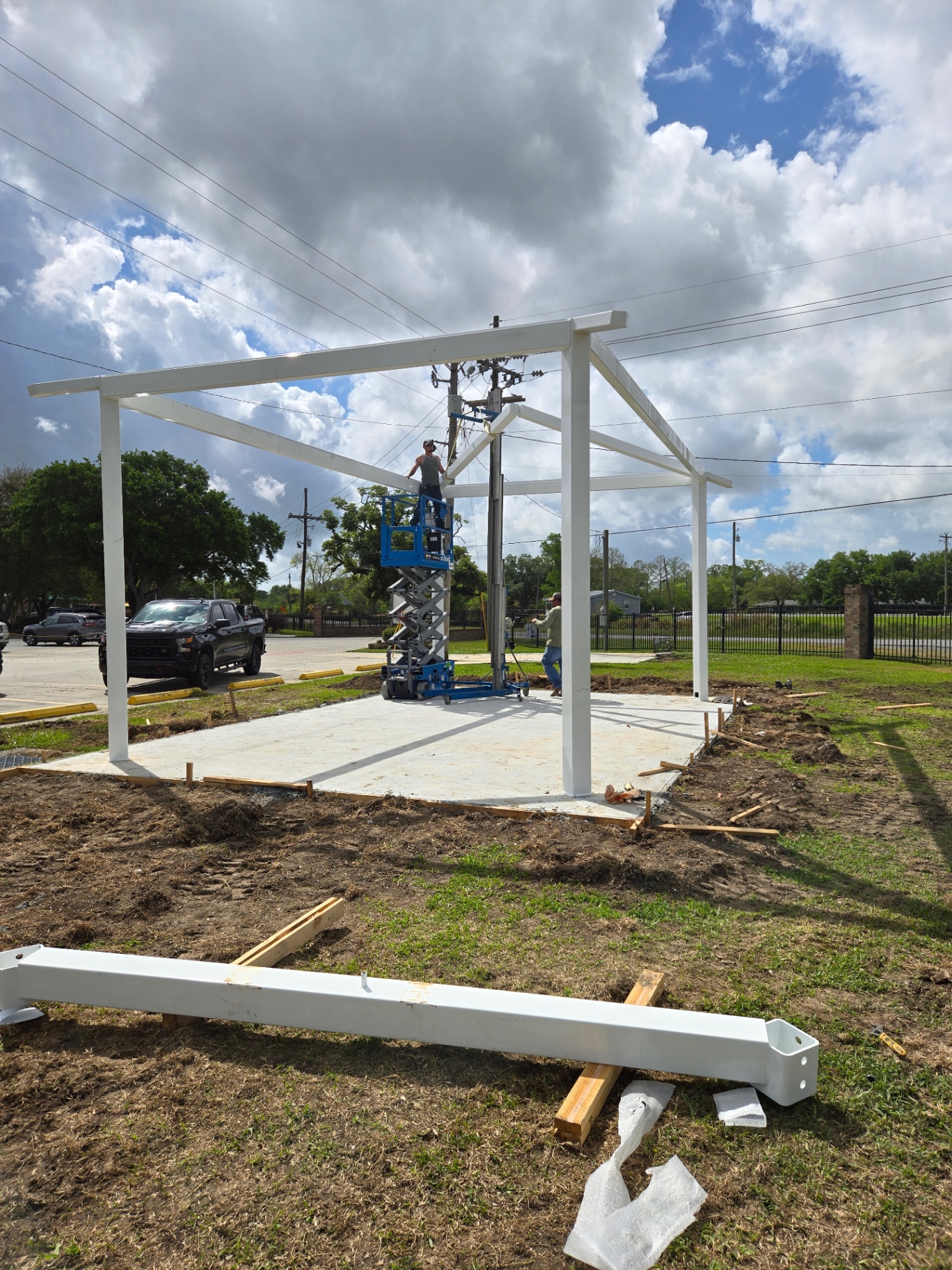 Welded steel pavilion frame being erected over a new concrete slab with a scissor lift on site